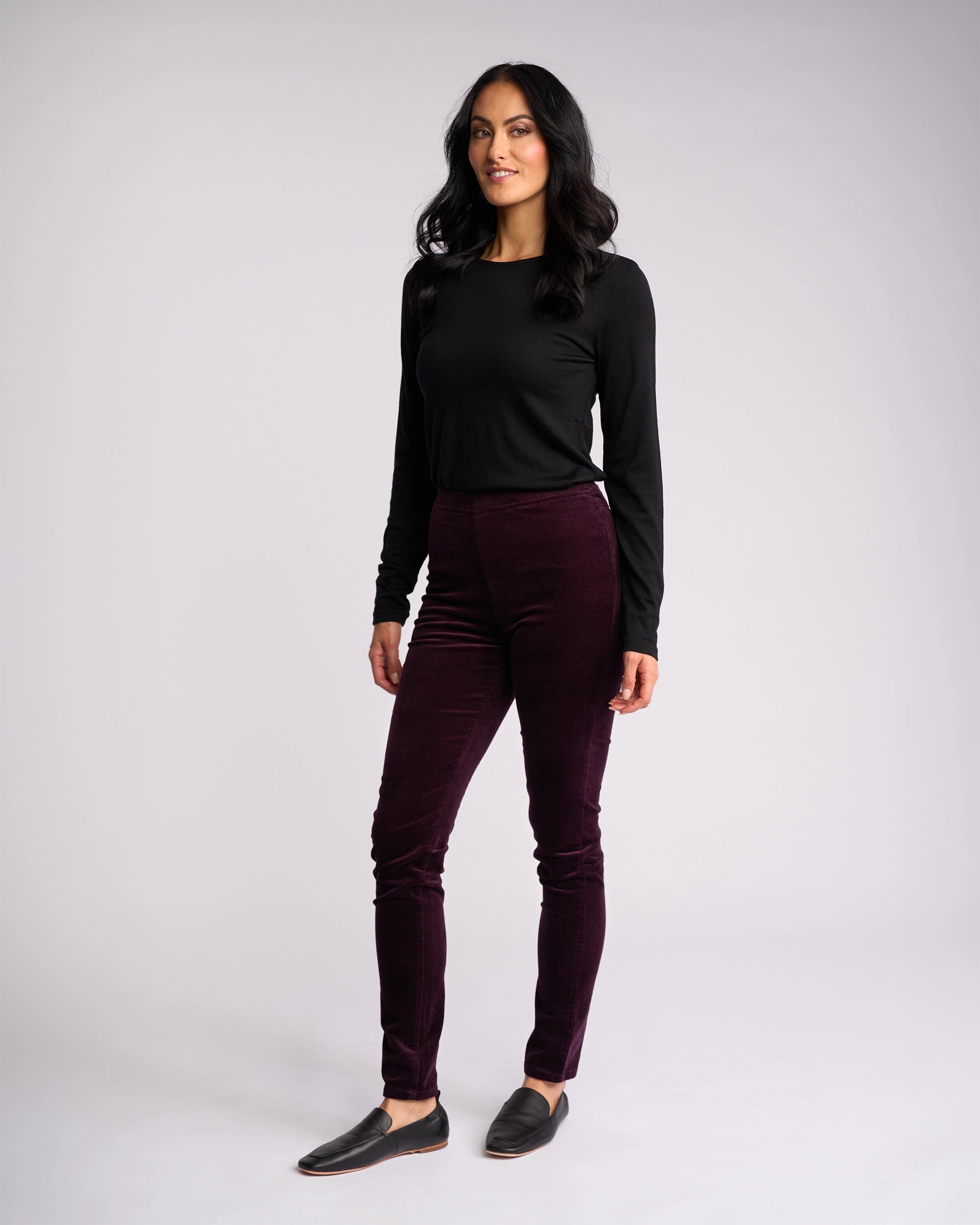 A woman with long dark hair poses against a plain background, wearing Vassalli’s Origin Cord Pull On pants in Mulberry with a black long-sleeve top and black slip-on shoes, smiling slightly as she stands with one leg forward.