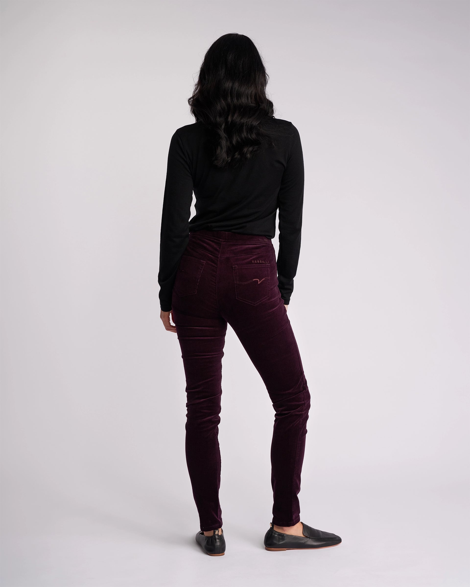 A person with long dark hair, wearing a black long-sleeve top and Vassalli’s Origin Cord Pull On - Mulberry skinny-leg pants, stands facing away from the camera against a plain light background.