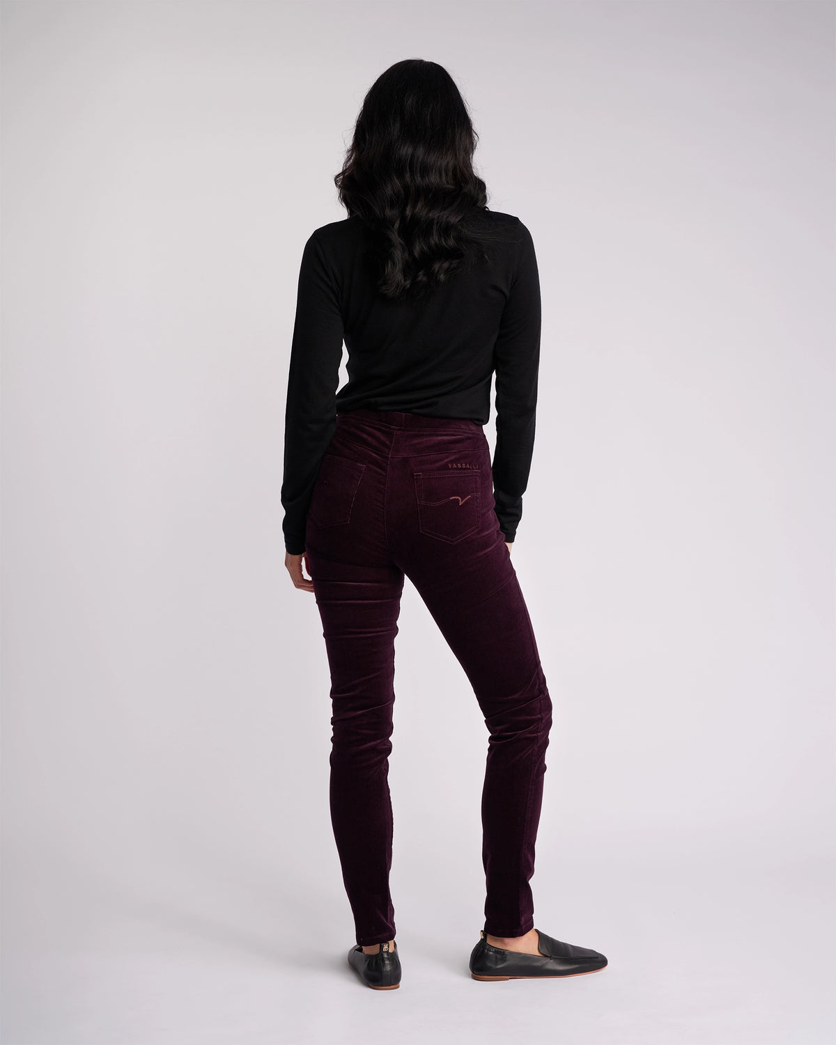 A person with long dark hair, wearing a black long-sleeve top and Vassalli’s Origin Cord Pull On - Mulberry skinny-leg pants, stands facing away from the camera against a plain light background.