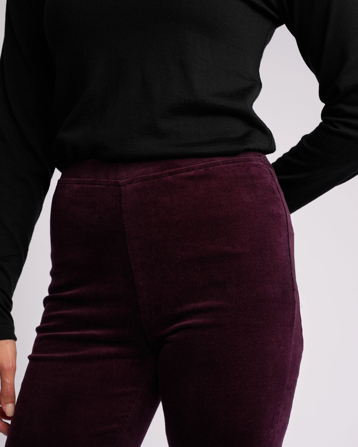 A person wearing a black long-sleeve top and Vassalli's Origin Cord Pull On skinny-leg pants in Mulberry, standing with one hand on their hip against a plain light background.