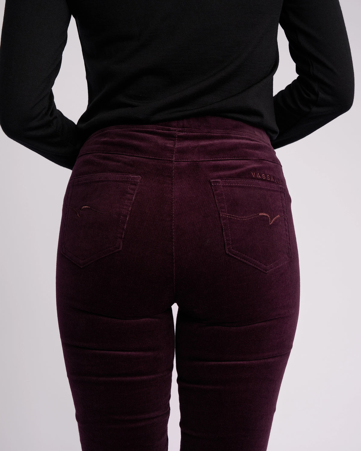 Model wears the Vassalli Origin Cord Pull On - Mulberry, a high-waisted, skinny-leg corduroy pant with a pull-on design, paired with a black long-sleeve top; shown from the back against a plain light background.