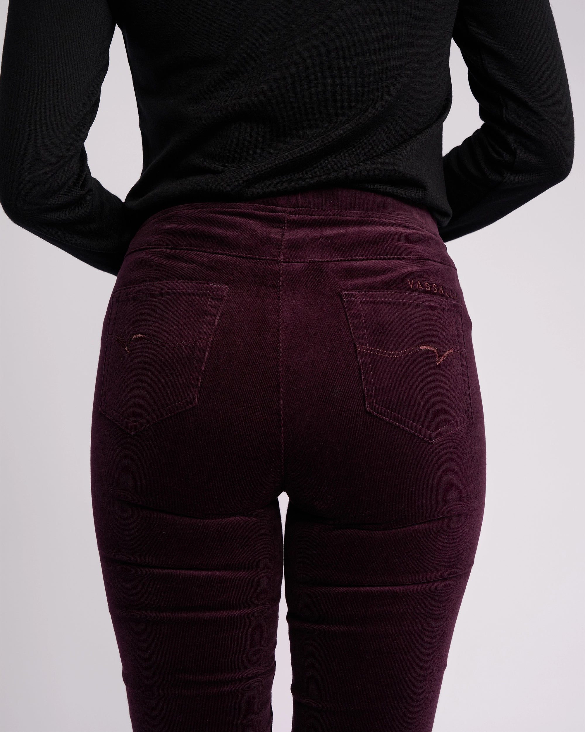 Model wears the Vassalli Origin Cord Pull On - Mulberry, a high-waisted, skinny-leg corduroy pant with a pull-on design, paired with a black long-sleeve top; shown from the back against a plain light background.