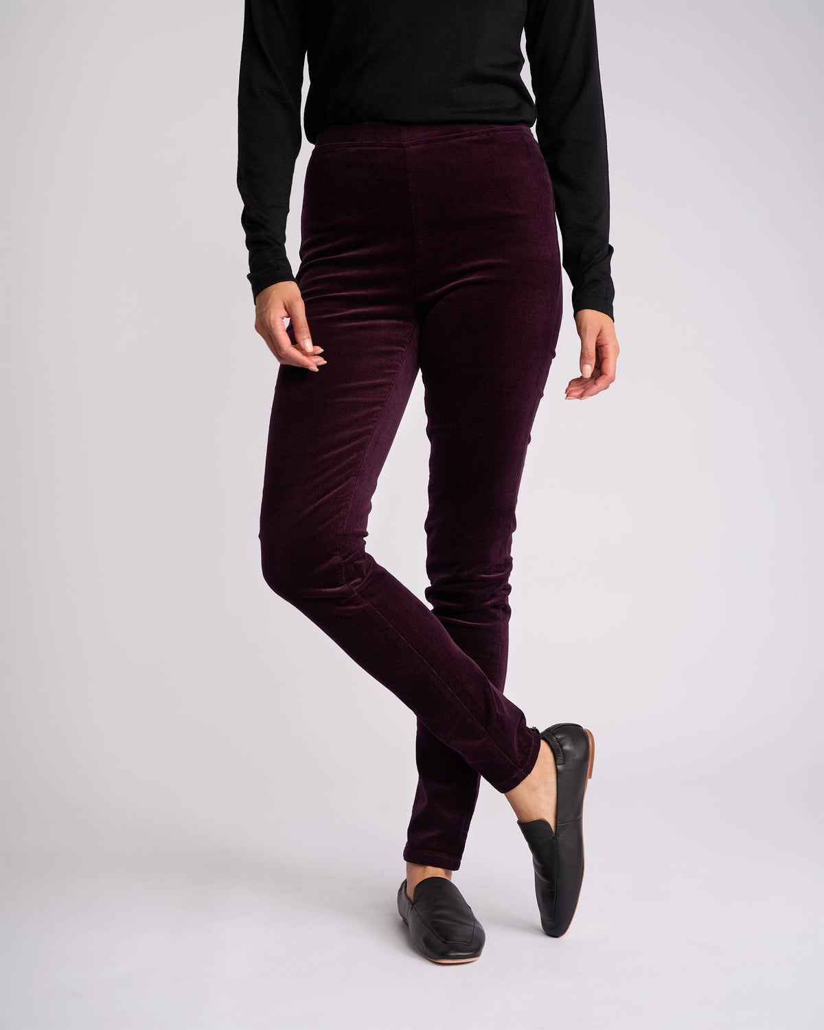 A person wears a black long-sleeve top, Vassalli Origin Cord Pull On pants in Mulberry, and black slip-on shoes, standing with one leg crossed in front of the other against a plain light background.