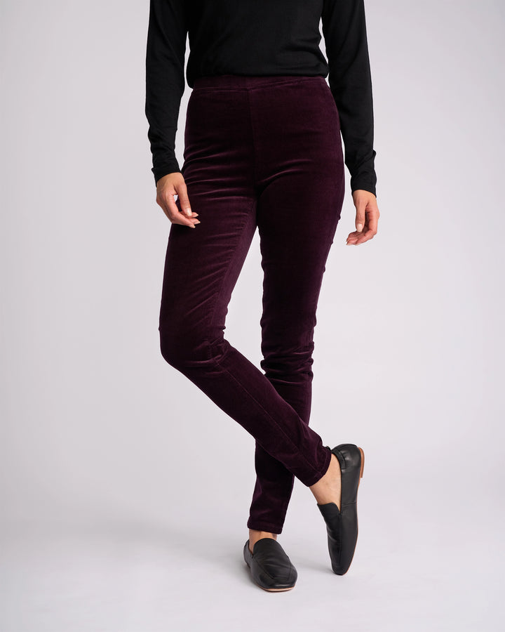 A person wears a black long-sleeve top, Vassalli Origin Cord Pull On pants in Mulberry, and black slip-on shoes, standing with one leg crossed in front of the other against a plain light background.