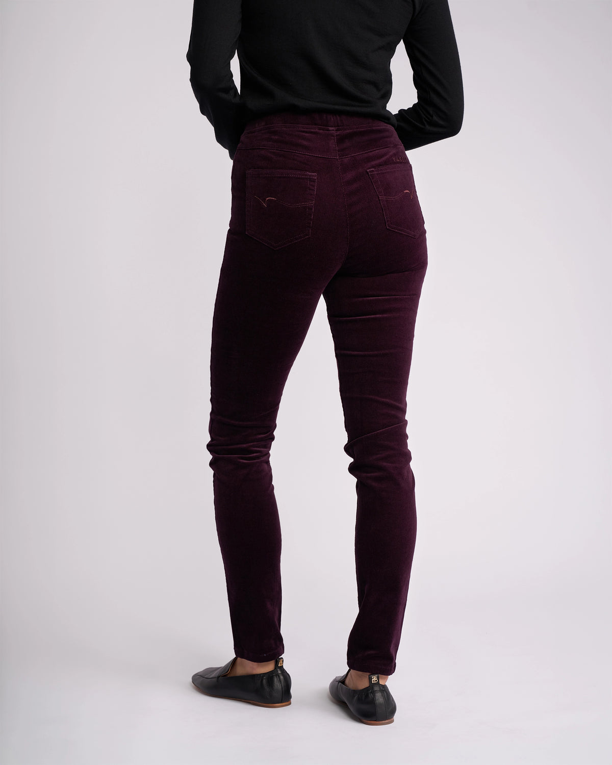 A person wearing a black long-sleeve top, Vassalli Origin Cord Pull On - Mulberry pants, and black flat shoes stands with their back to the camera against a plain light background.