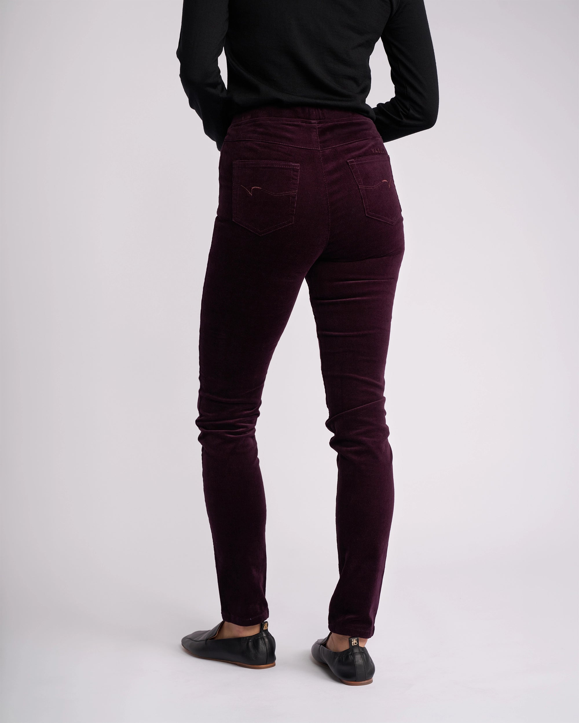 A person wearing a black long-sleeve top, Vassalli Origin Cord Pull On - Mulberry pants, and black flat shoes stands with their back to the camera against a plain light background.