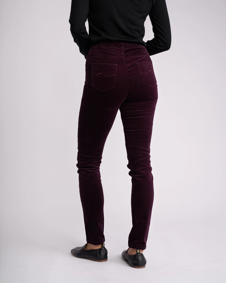 A person wearing a black long-sleeve top, Vassalli Origin Cord Pull On - Mulberry pants, and black flat shoes stands with their back to the camera against a plain light background.