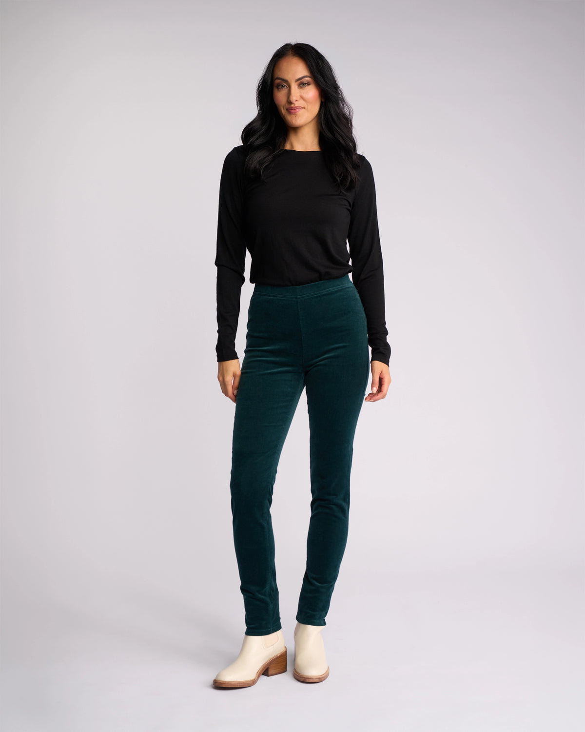 A woman with long dark hair and a confident look stands against a light gray background, wearing the Vassalli Origin Cord Pull On pants in Peacock, a long-sleeve black top, and cream ankle boots.