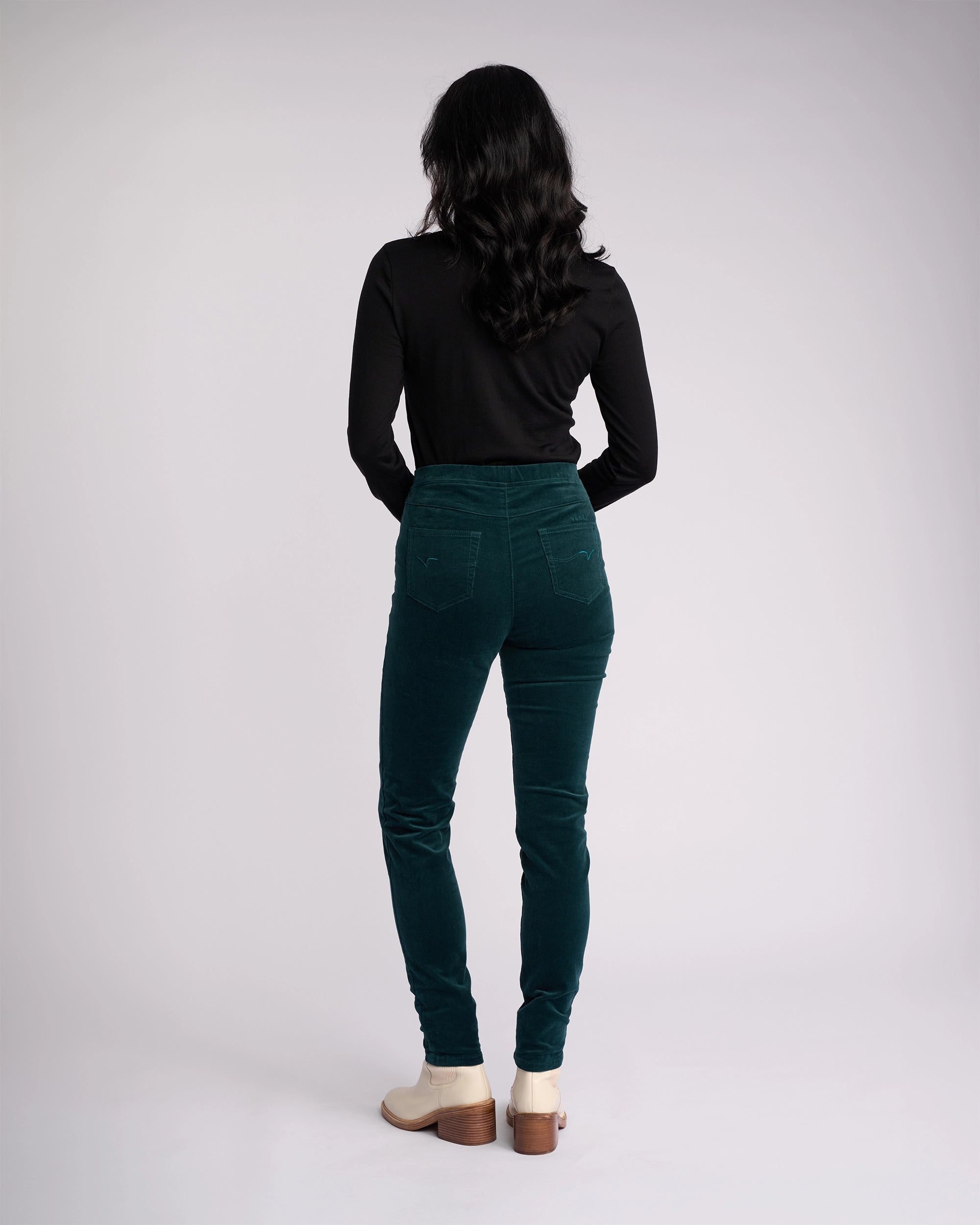 A woman with wavy dark hair stands facing away, wearing a black long-sleeve top and Vassalli Origin Cord Pull On pants in Peacock, paired with light-colored shoes, against a plain light background.