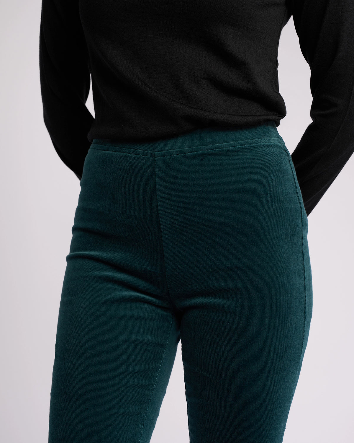 A person stands with arms behind their back, wearing a Vassalli long-sleeved black top and high-waisted Origin Cord Pull On - Peacock skinny-leg pants. The image is cropped from shoulders to upper thighs.
