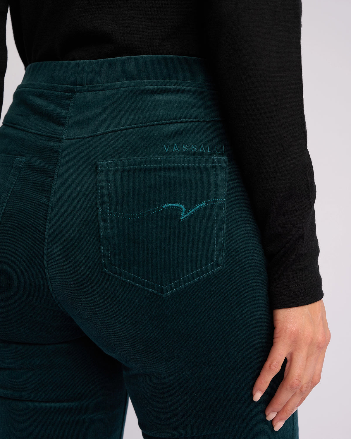 A person wears Vassalli Origin Cord Pull On pants in Peacock, paired with a black long-sleeve top. The skinny-leg, stretch pinwale cord pant features a pull-on design, minimal back pocket stitching, and "VASSALLI" embroidered above the pocket.
