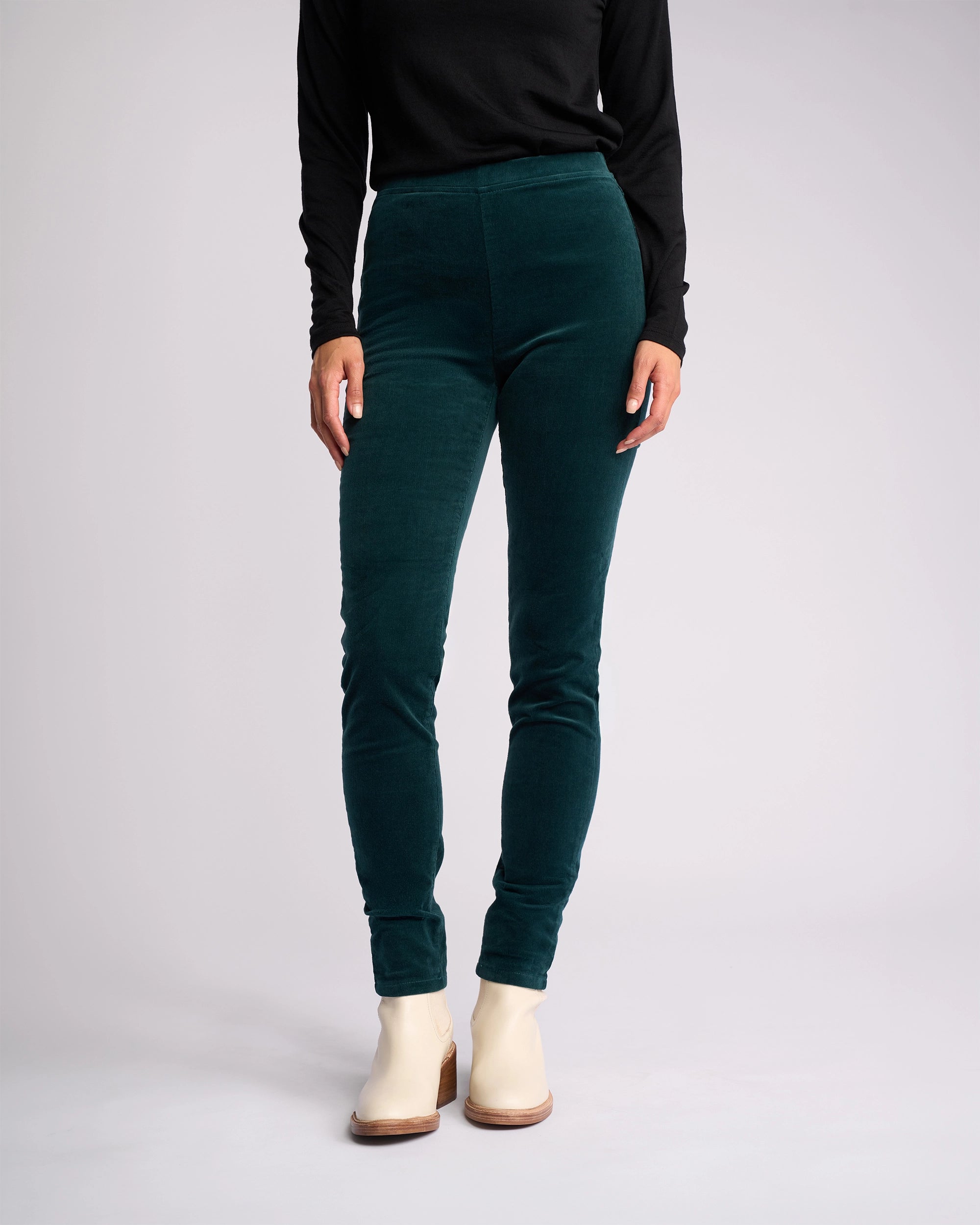 A person models Vassalli's Origin Cord Pull On - Peacock pants with a long-sleeve black top and white ankle boots against a plain light background. Their face is not shown.