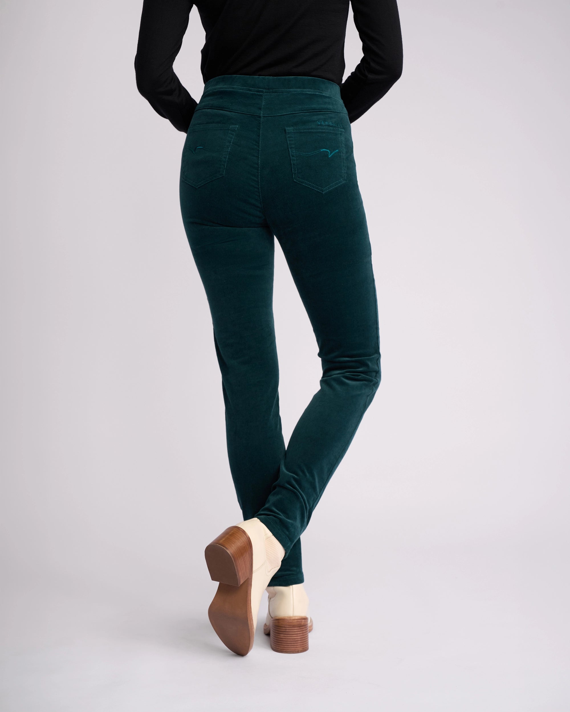A person wearing Vassalli Origin Cord Pull On pants in Peacock, a long-sleeve black top, and beige heeled shoes stands with their back to the camera against a plain light background.