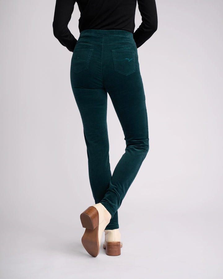 A person wearing Vassalli Origin Cord Pull On pants in Peacock, a long-sleeve black top, and beige heeled shoes stands with their back to the camera against a plain light background.