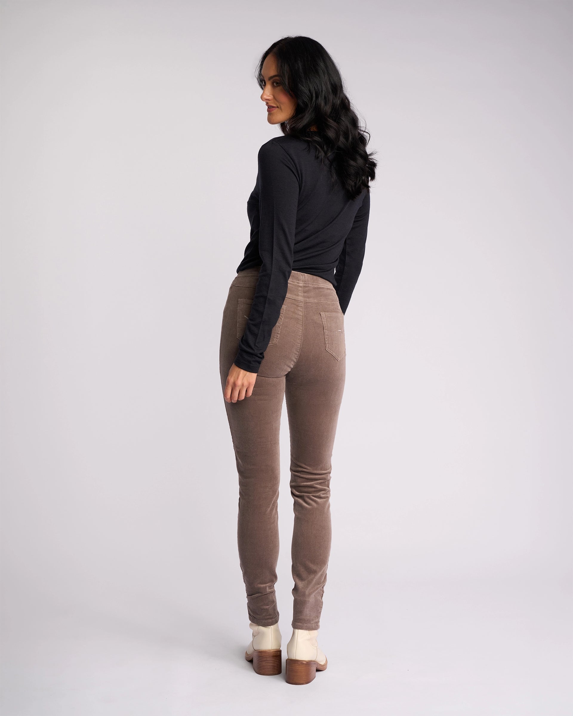 A woman with long dark hair faces away, turning her head sideward. She wears a black long-sleeve top, beige-heeled shoes, and Vassalli Origin Cord Pull On pants in taupe against a plain light background.