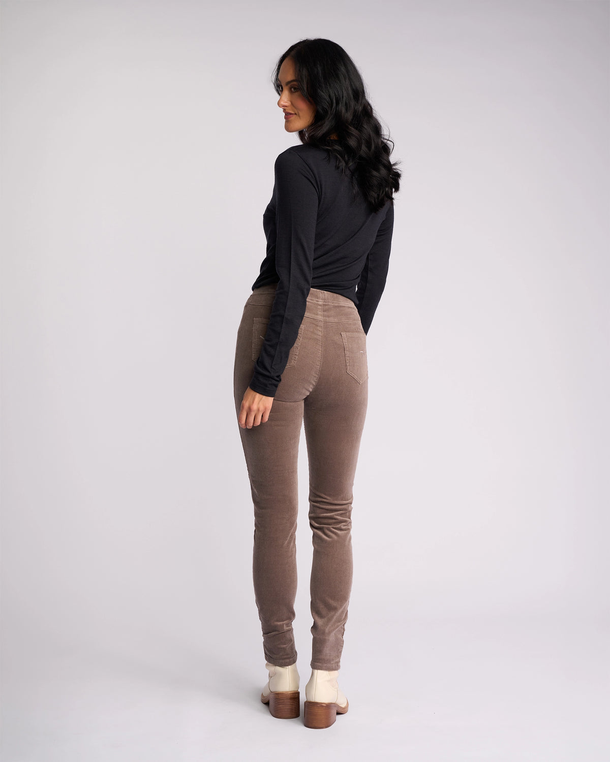 A woman with long dark hair faces away, turning her head sideward. She wears a black long-sleeve top, beige-heeled shoes, and Vassalli Origin Cord Pull On pants in taupe against a plain light background.