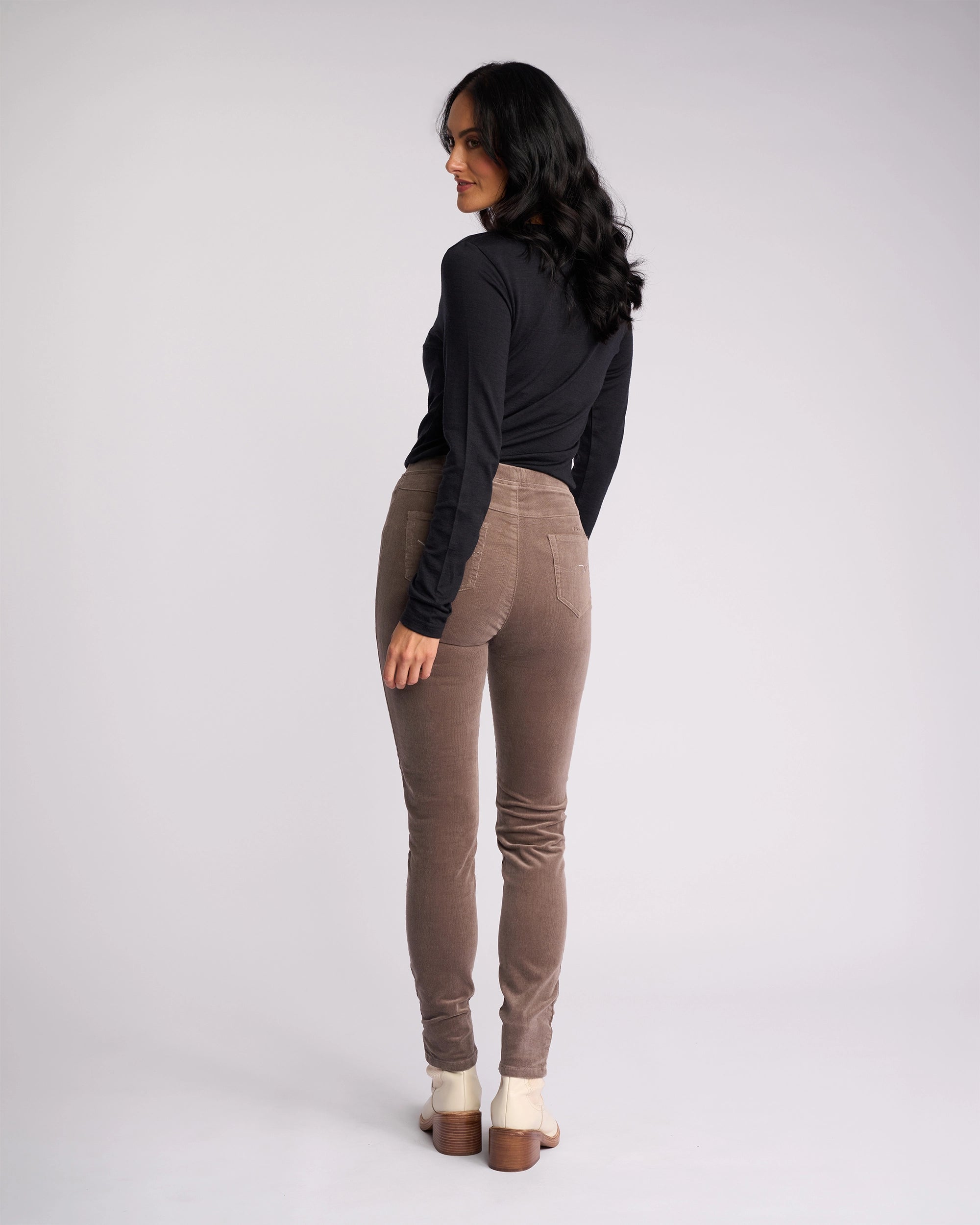 A woman with long dark hair faces away, turning her head sideward. She wears a black long-sleeve top, beige-heeled shoes, and Vassalli Origin Cord Pull On pants in taupe against a plain light background.
