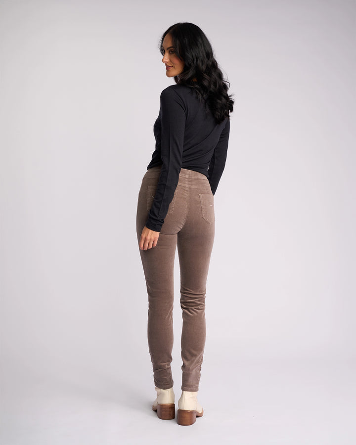 A woman with long dark hair faces away, turning her head sideward. She wears a black long-sleeve top, beige-heeled shoes, and Vassalli Origin Cord Pull On pants in taupe against a plain light background.