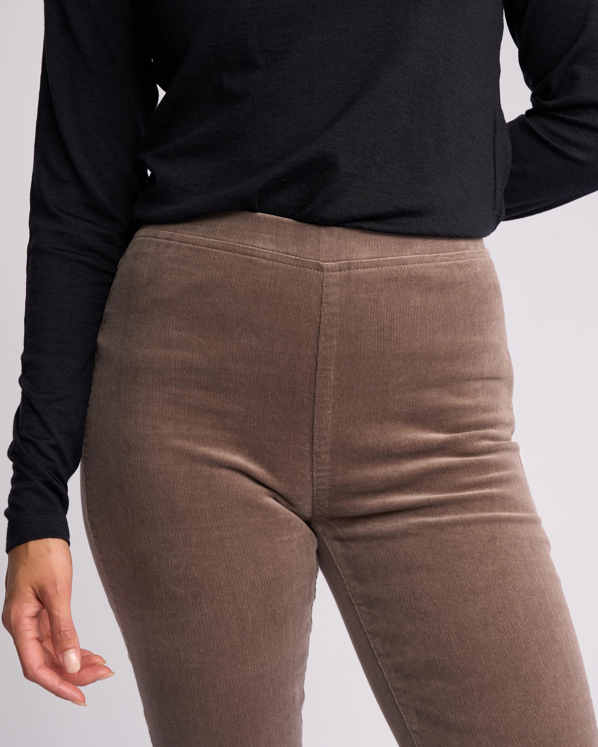 A person wearing a long-sleeve black top and Vassalli Origin Cord Pull On pants in taupe stands against a light background, one arm slightly bent and the other by their side.