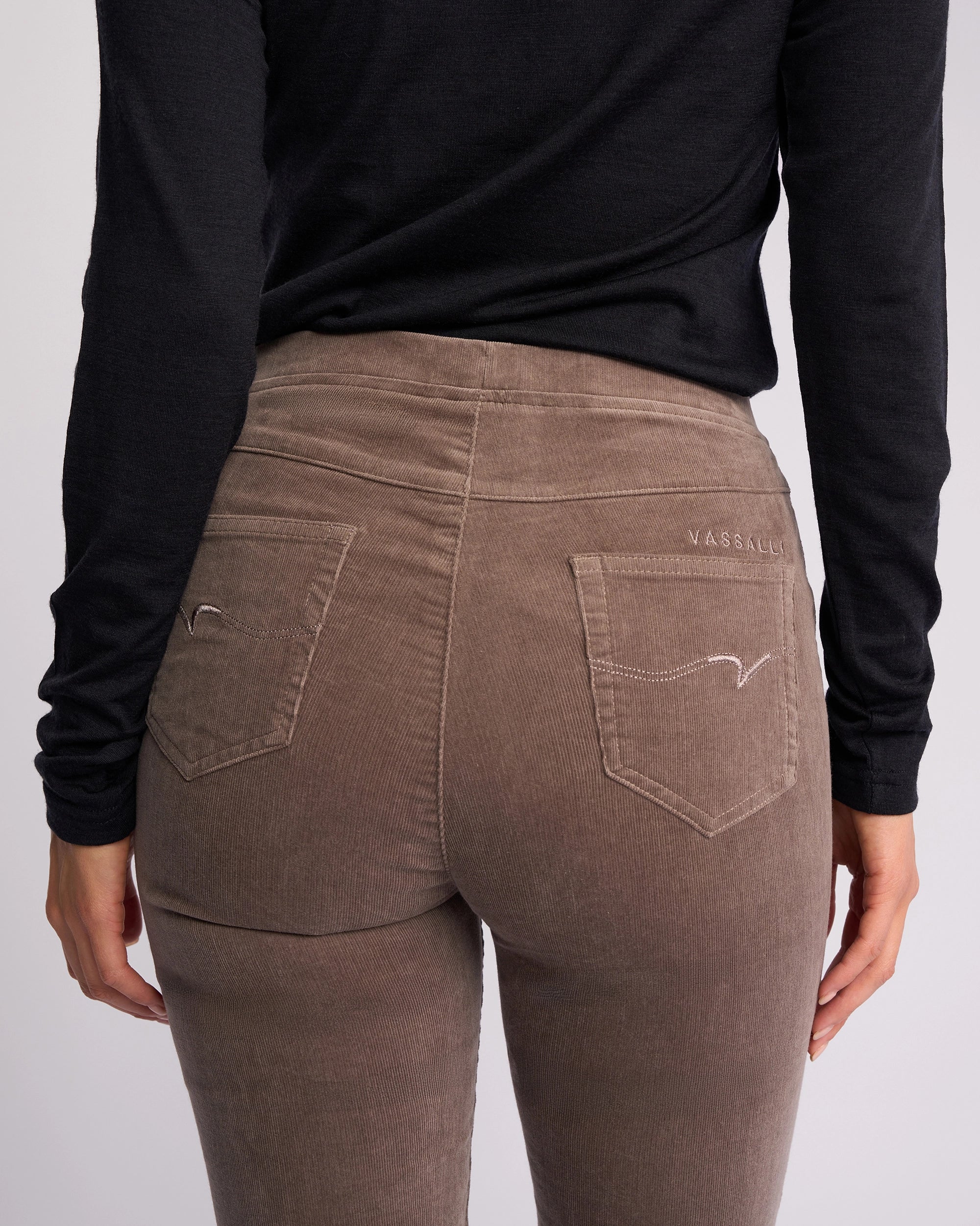 A person is shown from the back wearing a long-sleeve black top and Vassalli Origin Cord Pull On - Taupe skinny-leg pants, featuring visible back pockets and stitched details.