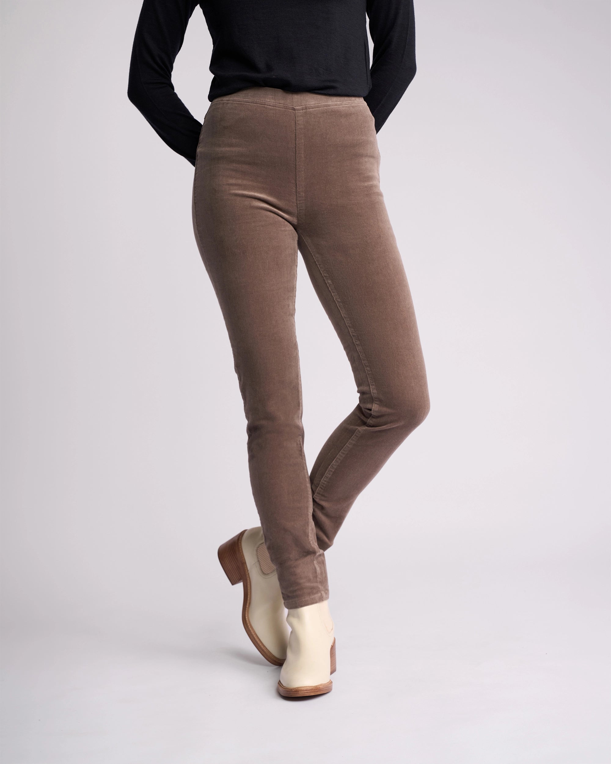 A person, face not visible, stands against a plain background wearing a black long-sleeve top, Vassalli Origin Cord Pull On pants in taupe, and light beige ankle boots with their hands behind their back.