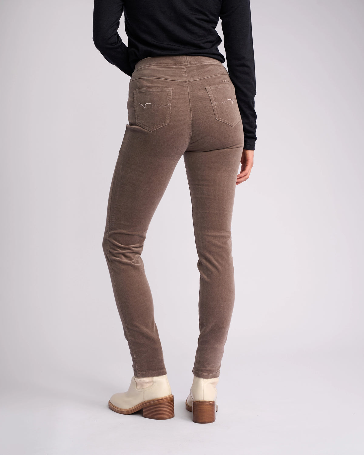 A person wearing Vassalli’s Origin Cord Pull On pants in taupe, paired with a black long-sleeve top and off-white heeled ankle boots, stands facing away against a plain light background.