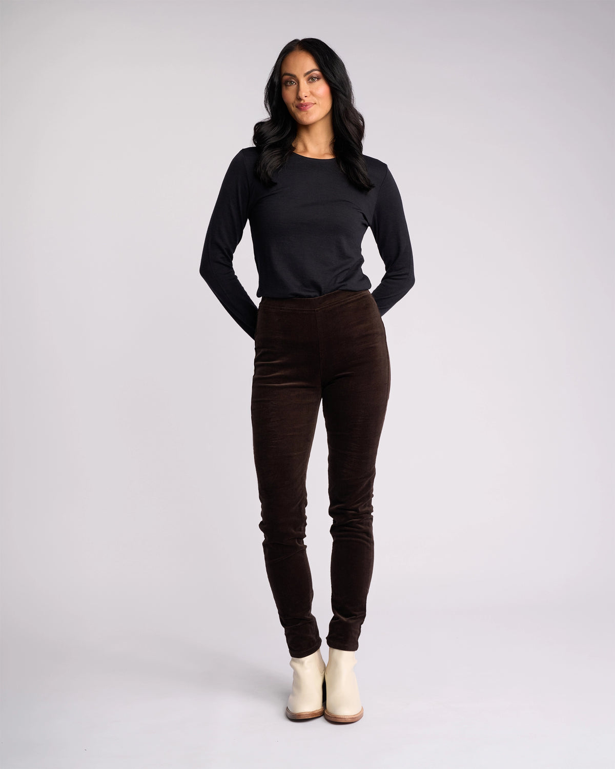 A woman with long dark hair wears a black long-sleeve top, Vassalli Origin Cord Pull On pants in Walnut, and light ankle boots. She stands facing forward with her hands behind her back against a plain light background.