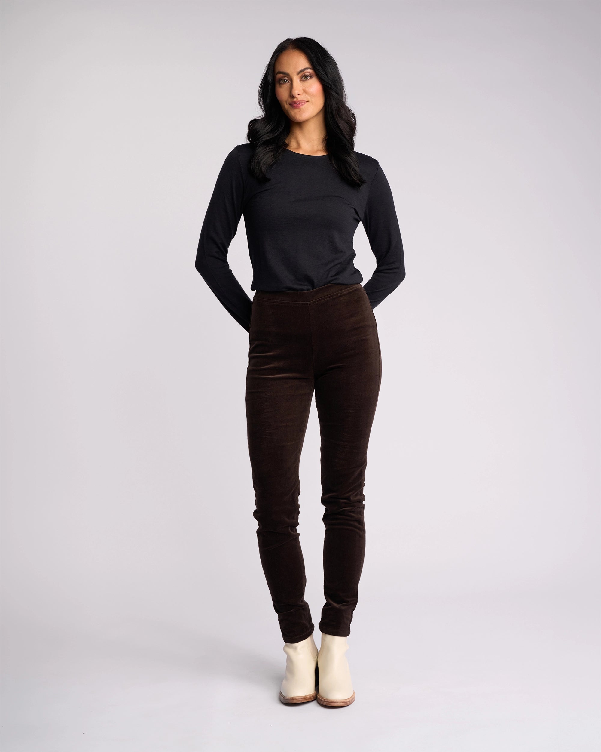 A woman with long dark hair wears a black long-sleeve top, Vassalli Origin Cord Pull On pants in Walnut, and light ankle boots. She stands facing forward with her hands behind her back against a plain light background.