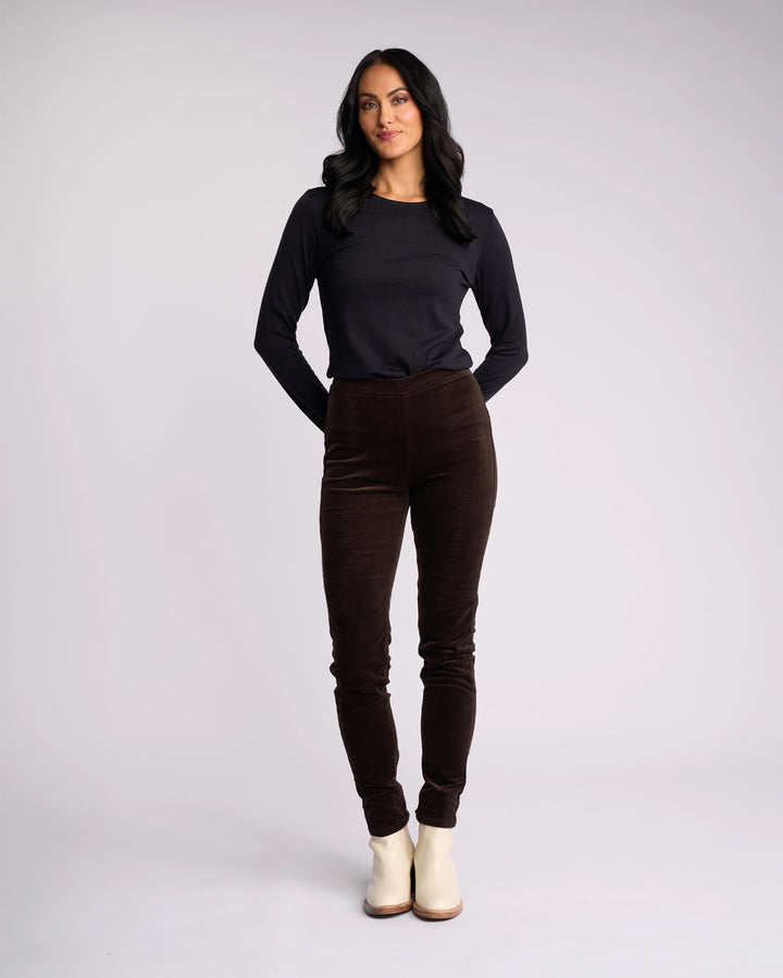 A woman with long dark hair wears a black long-sleeve top, Vassalli Origin Cord Pull On pants in Walnut, and light ankle boots. She stands facing forward with her hands behind her back against a plain light background.
