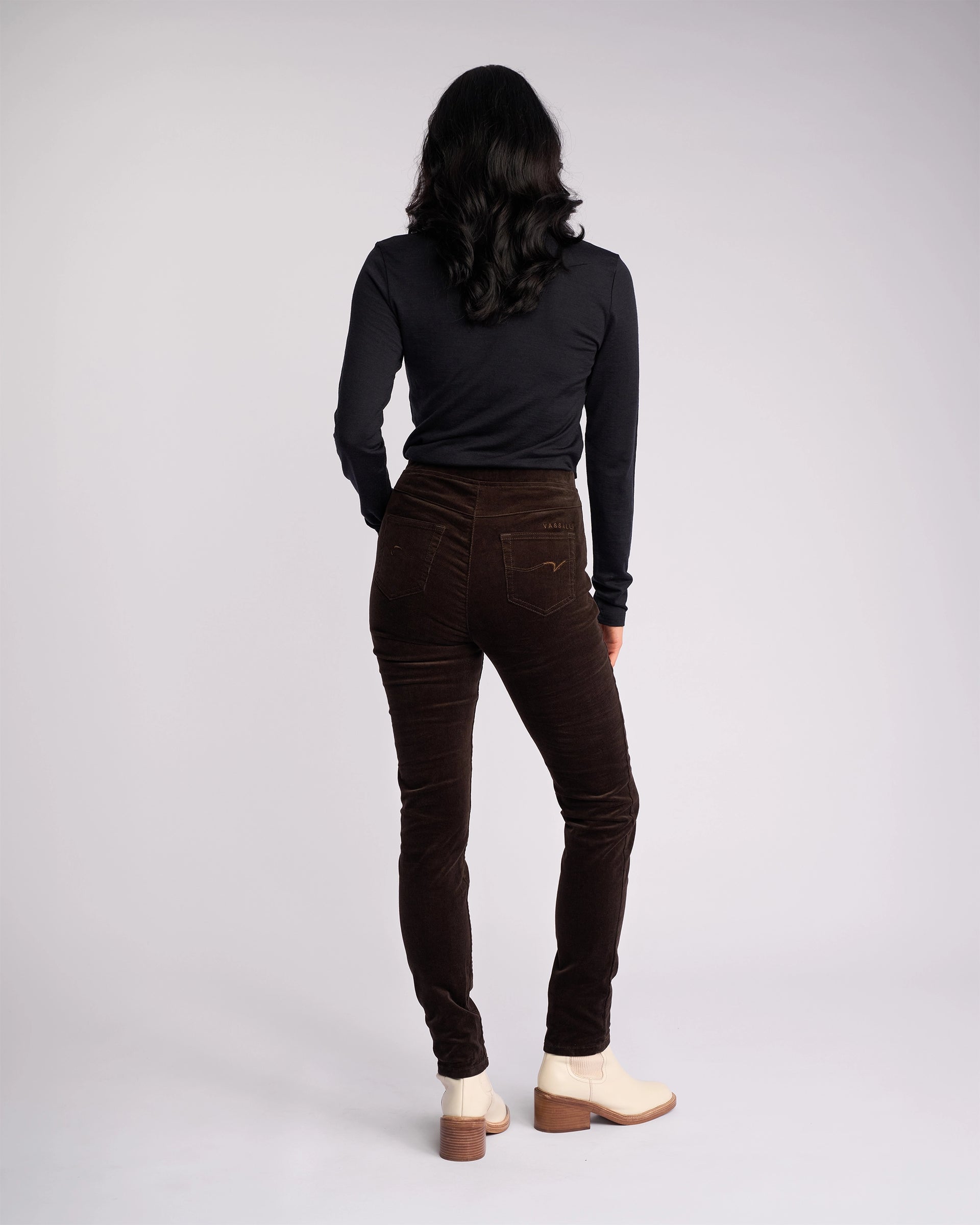 A person with long dark hair, wearing a black long-sleeve top and Vassalli Origin Cord Pull On - Walnut pants, stands facing away from the camera against a plain light background.