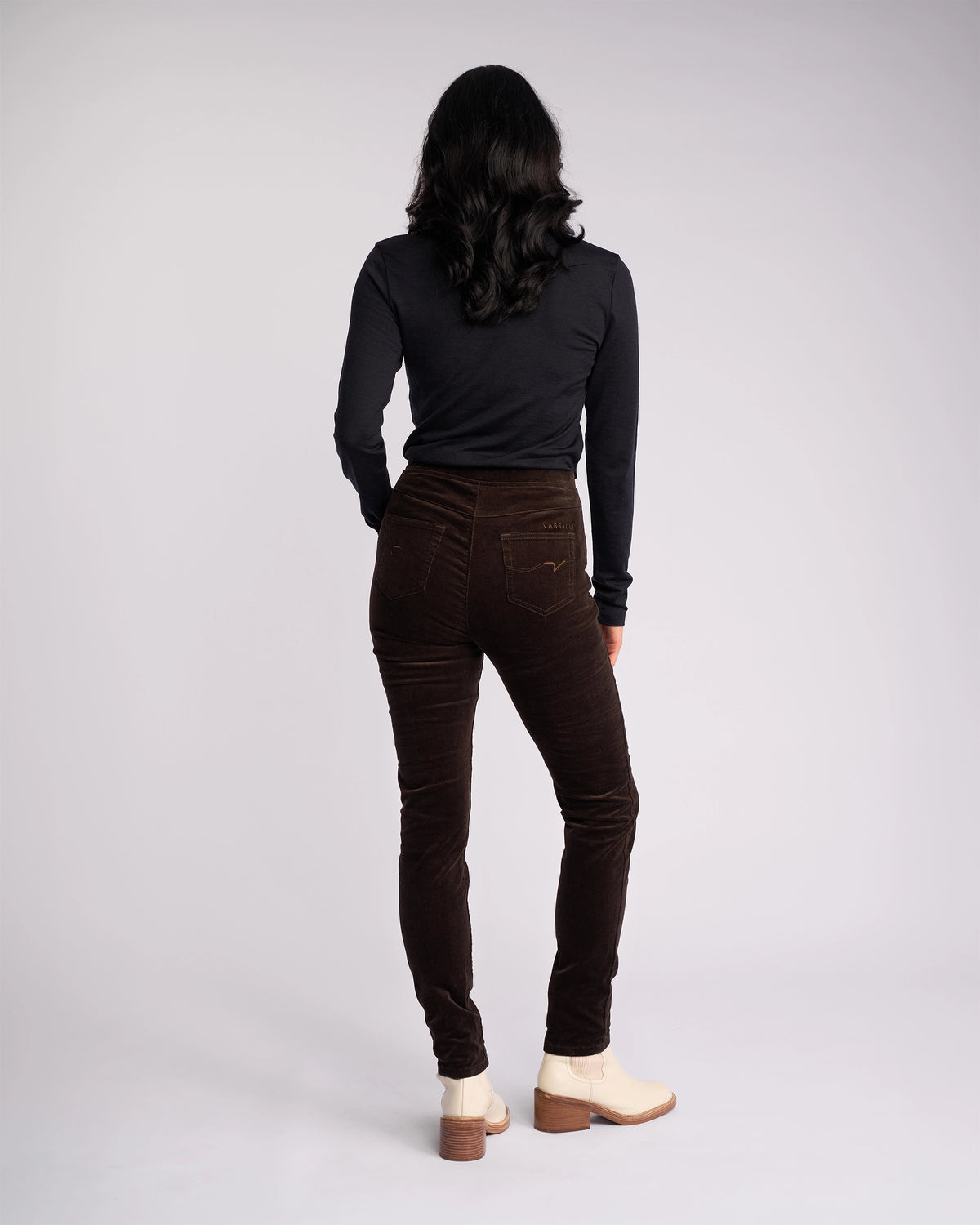 A person with long dark hair, wearing a black long-sleeve top and Vassalli Origin Cord Pull On - Walnut pants, stands facing away from the camera against a plain light background.