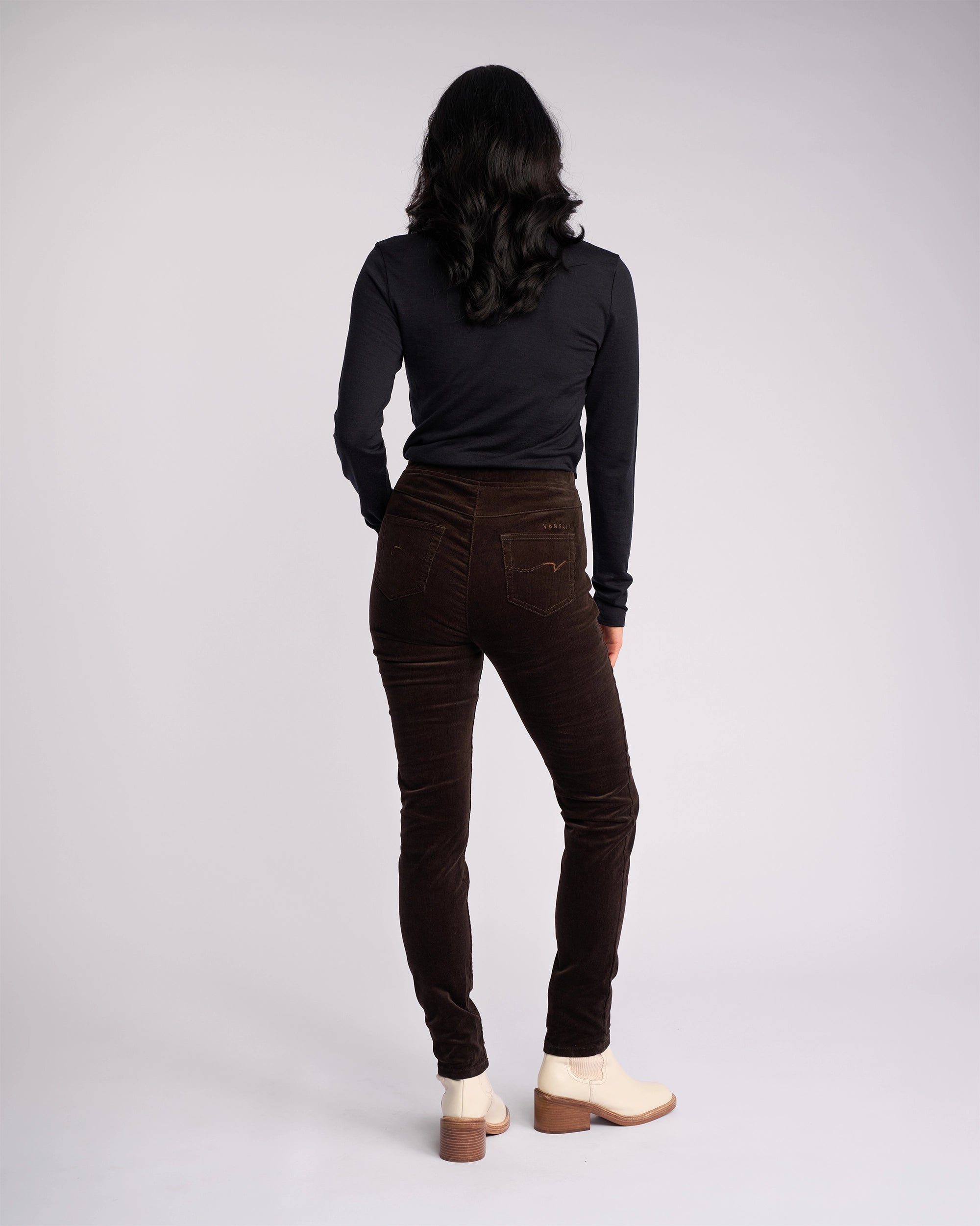 A person with long dark hair, wearing a black long-sleeve top and Vassalli Origin Cord Pull On - Walnut pants, stands facing away from the camera against a plain light background.
