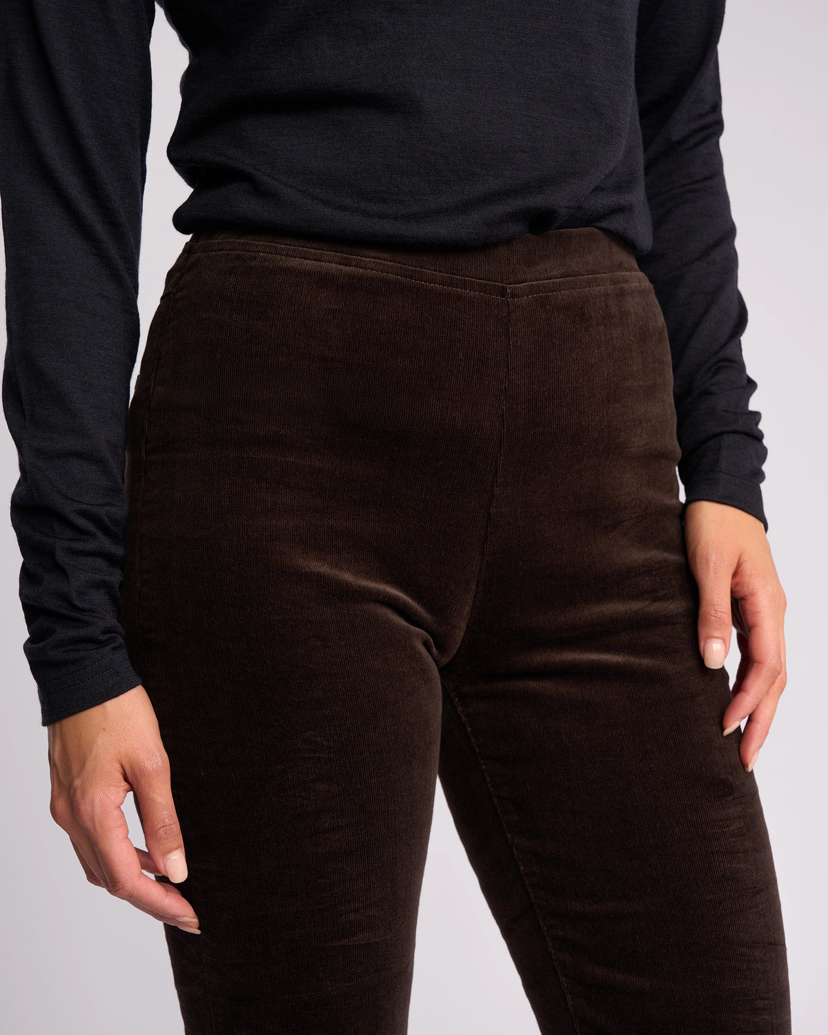 Someone stands against a plain light background wearing Vassalli’s Origin Cord Pull On pants in walnut, paired with a long-sleeve black top, with one hand resting by their side.