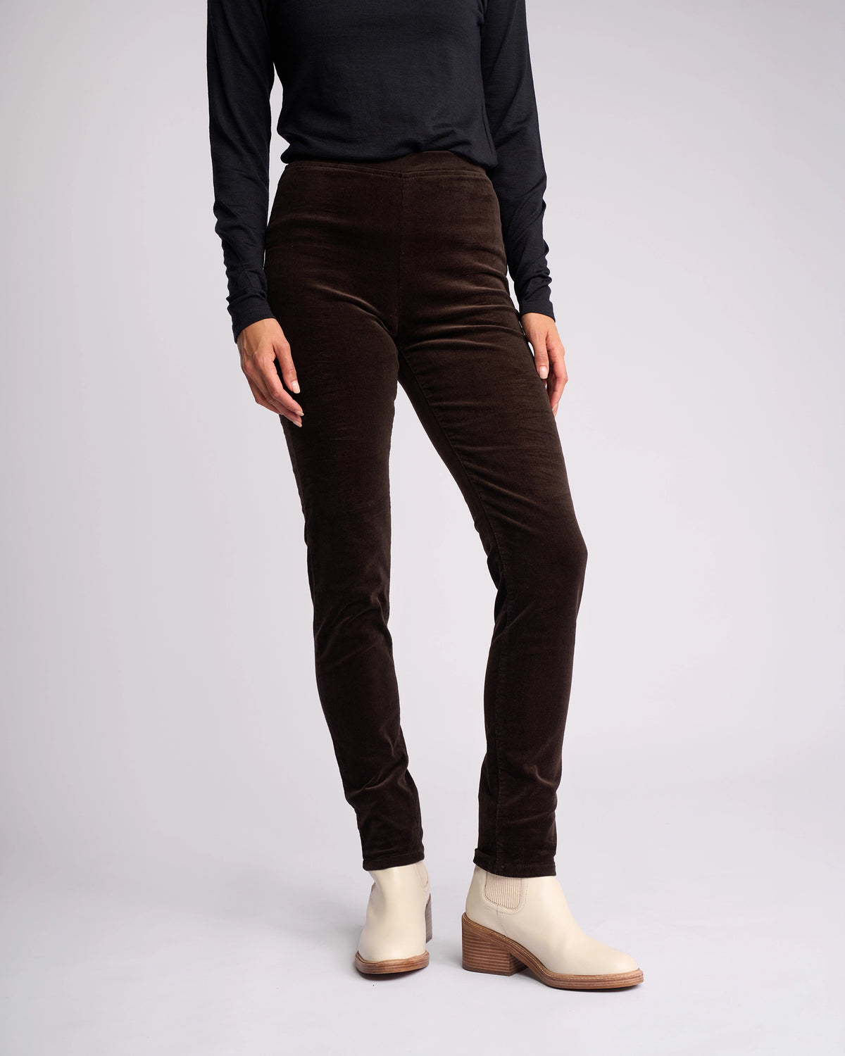 Wearing a long-sleeve black top, Vassalli Origin Cord Pull On - Walnut skinny pants, and light beige ankle boots with a wooden heel, a person stands against a plain light background. Their head is not visible.