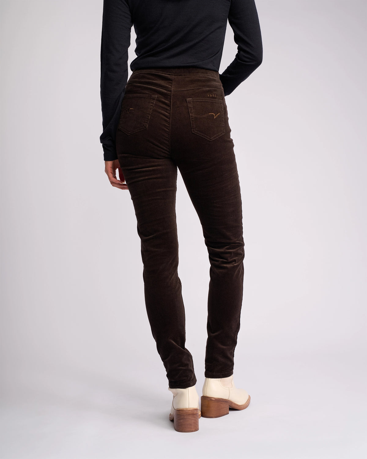 A person stands with their back to the camera against a plain, light background, wearing Vassalli Origin Cord Pull On pants in Walnut, paired with a black long-sleeve top and cream ankle boots.