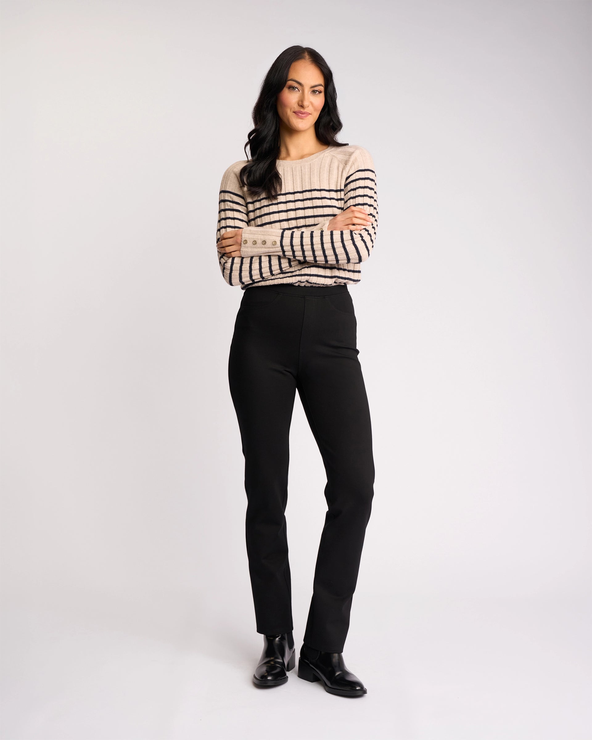 A woman with long dark hair stands with arms crossed, wearing a beige and black striped sweater, Vassalli Bay Ponti Pull On pants in black, and black shoes, against a plain light background.