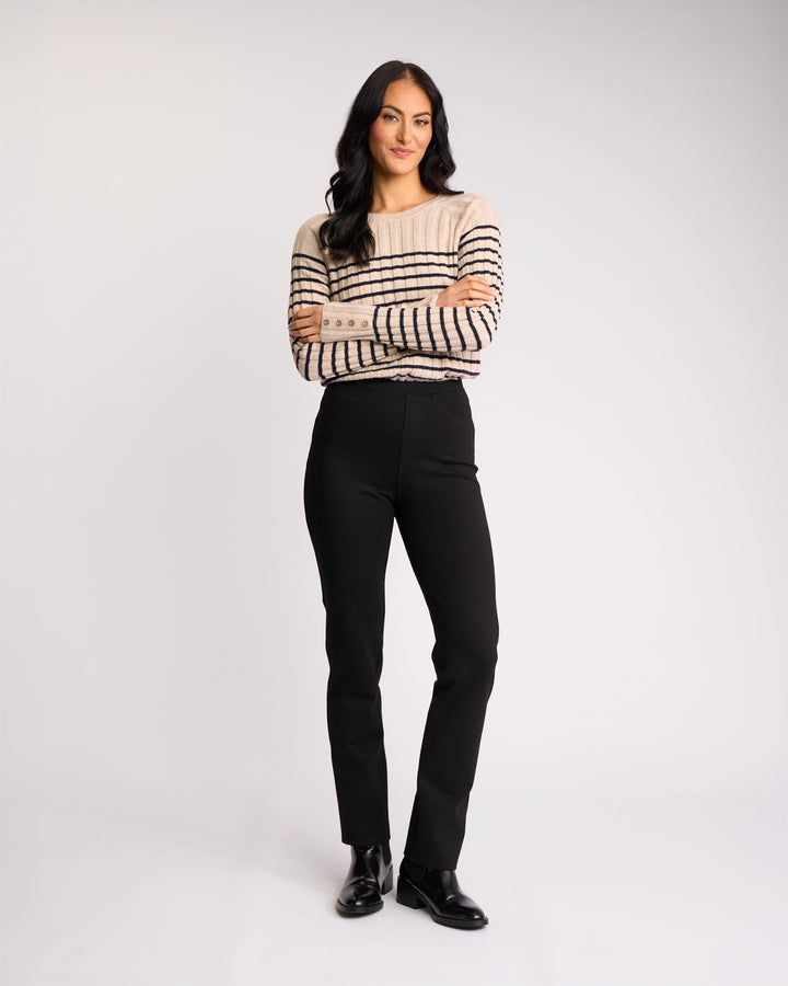 A woman with long dark hair stands with arms crossed, wearing a beige and black striped sweater, Vassalli Bay Ponti Pull On pants in black, and black shoes, against a plain light background.