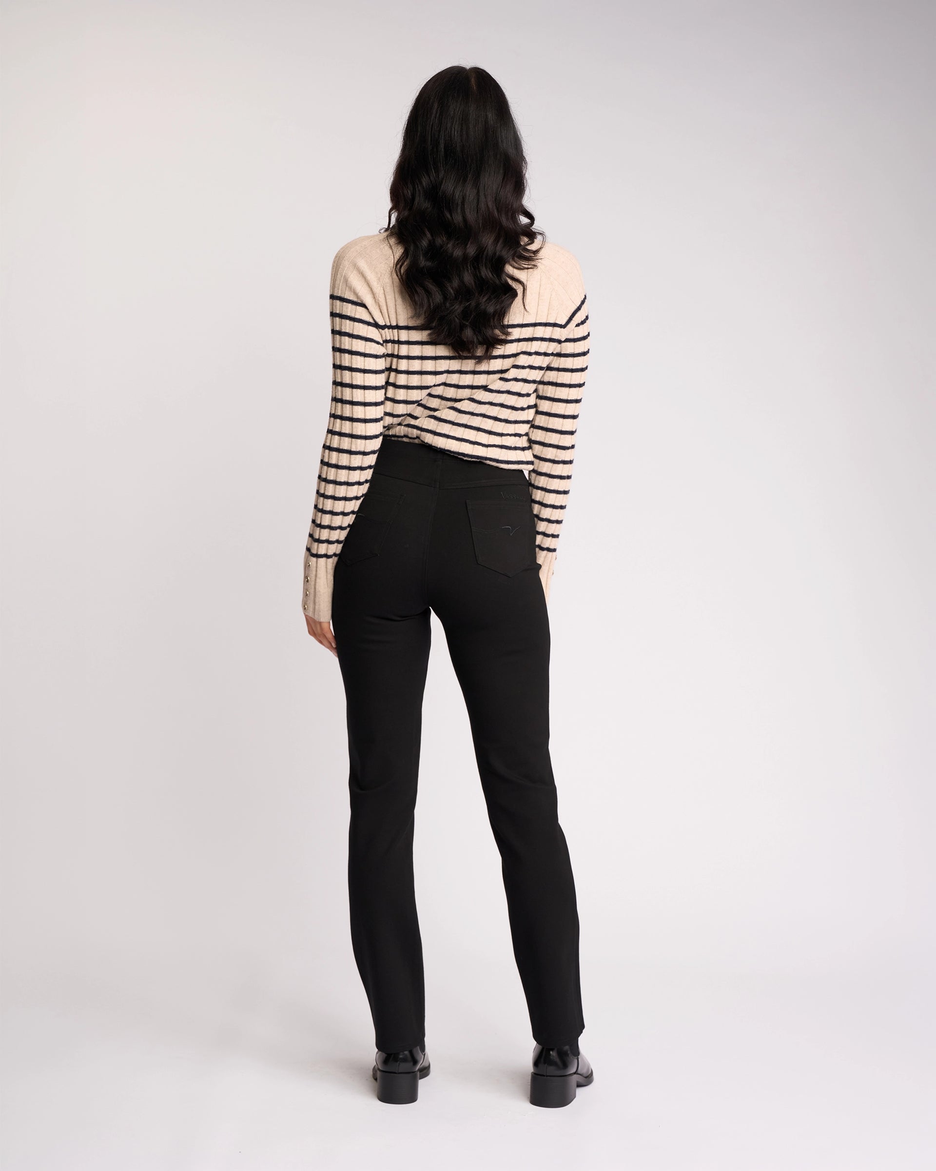 A person with long dark hair stands facing away, wearing a cream sweater with black stripes, Vassalli Bay Ponti Pull On pants in black, and black shoes against a plain light background.