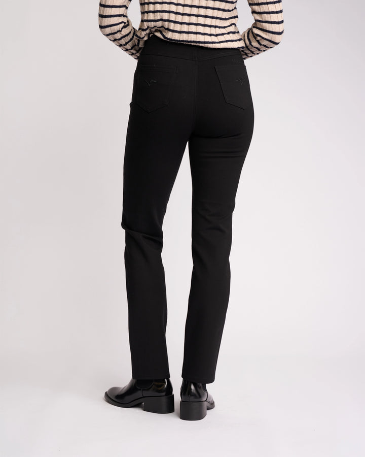 A person stands facing away from the camera, wearing Vassalli Bay Ponti Pull On pants in black, black shoes, and a long-sleeve striped beige and black top—versatile workwear styled against a plain light background.