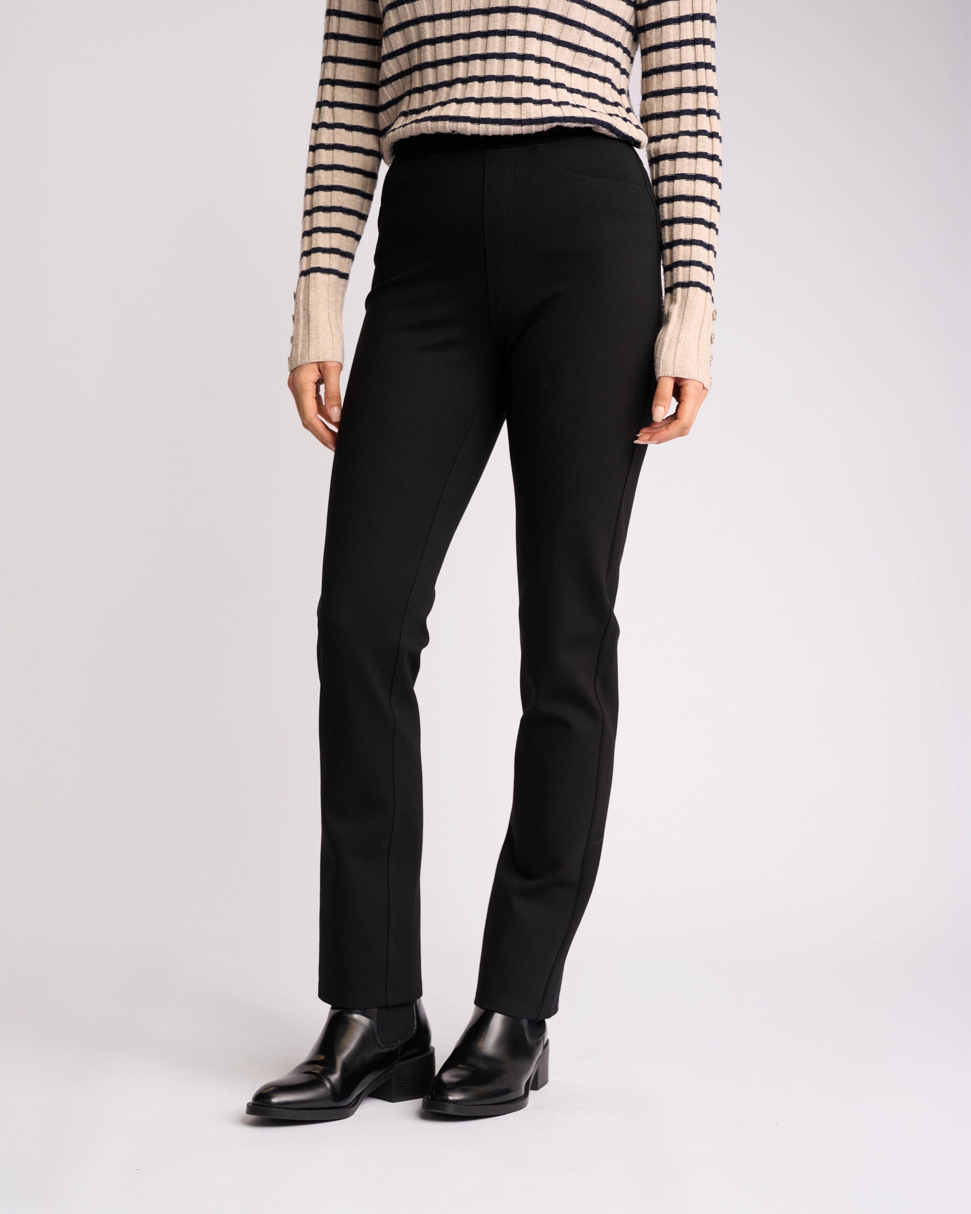 A person wearing Vassalli’s Bay Ponti Pull On - Black pants stands against a plain light background, paired with a beige and black striped sweater and black ankle boots. The image is cropped at the shoulders.
