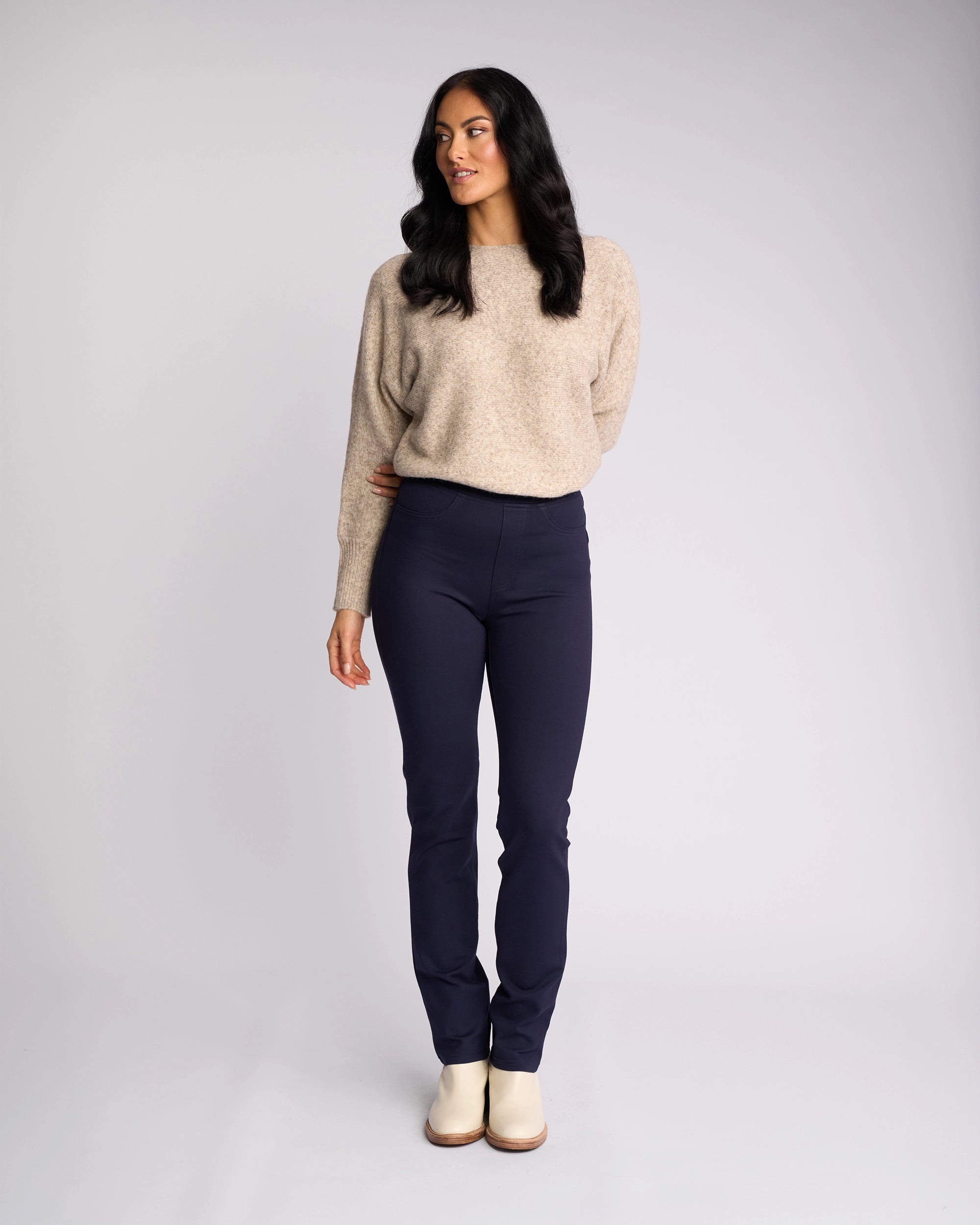 A woman with long dark hair stands against a light gray background, wearing the Vassalli Bay Ponti Pull On - Navy pants, a loose beige sweater, and cream shoes. She looks off to the side with relaxed arms.