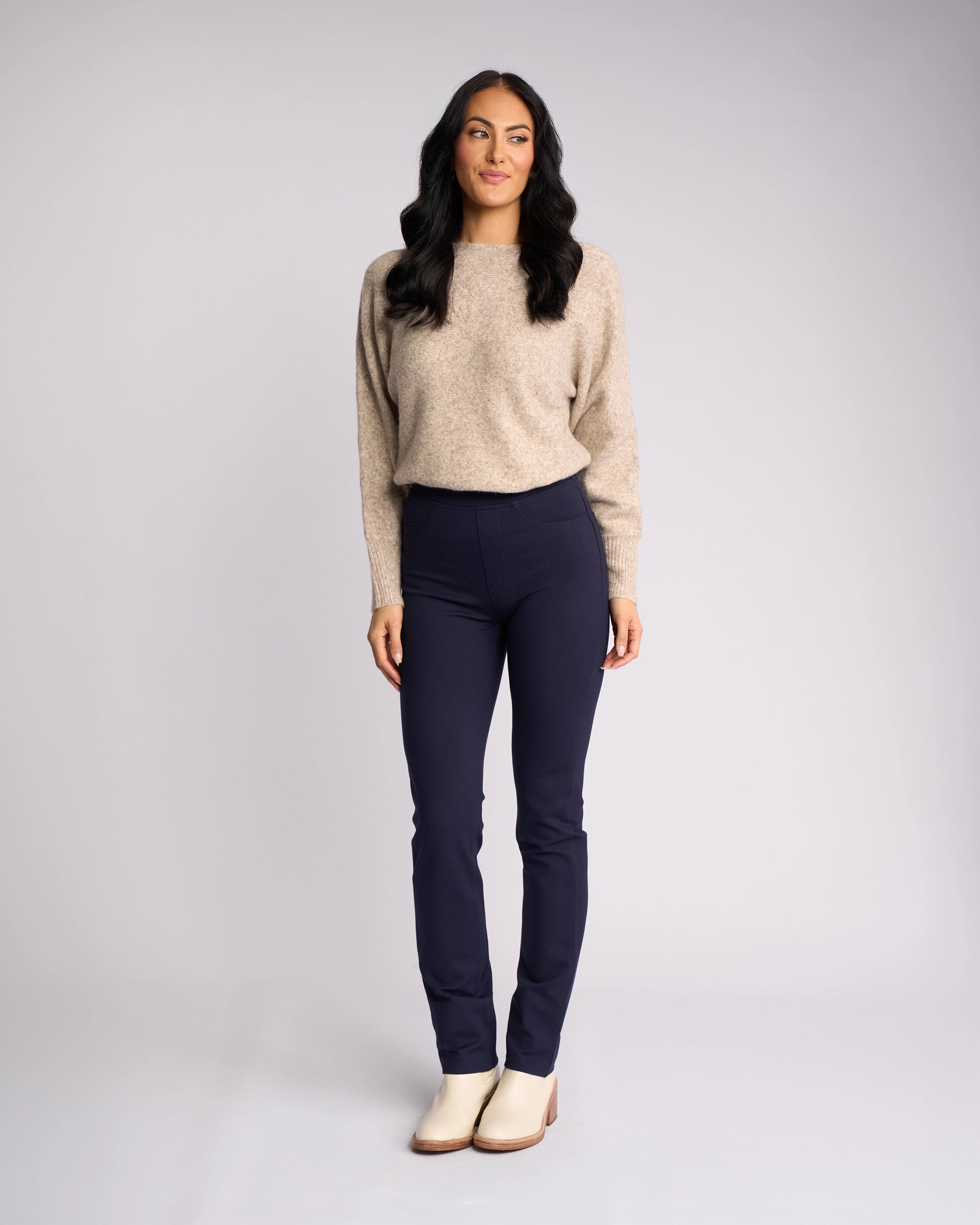 A woman with long dark hair stands against a neutral background, smiling slightly. She wears the Vassalli Bay Ponti Pull On - Navy pants with a beige sweater—ideal for modern workwear—her arms relaxed by her sides.