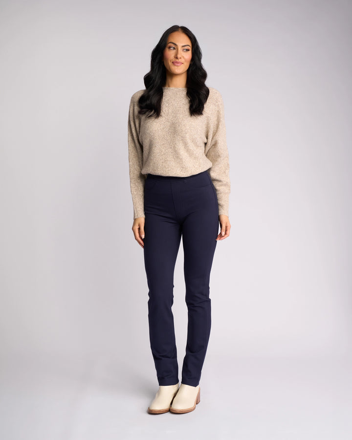 A woman with long dark hair stands against a neutral background, smiling slightly. She wears the Vassalli Bay Ponti Pull On - Navy pants with a beige sweater—ideal for modern workwear—her arms relaxed by her sides.