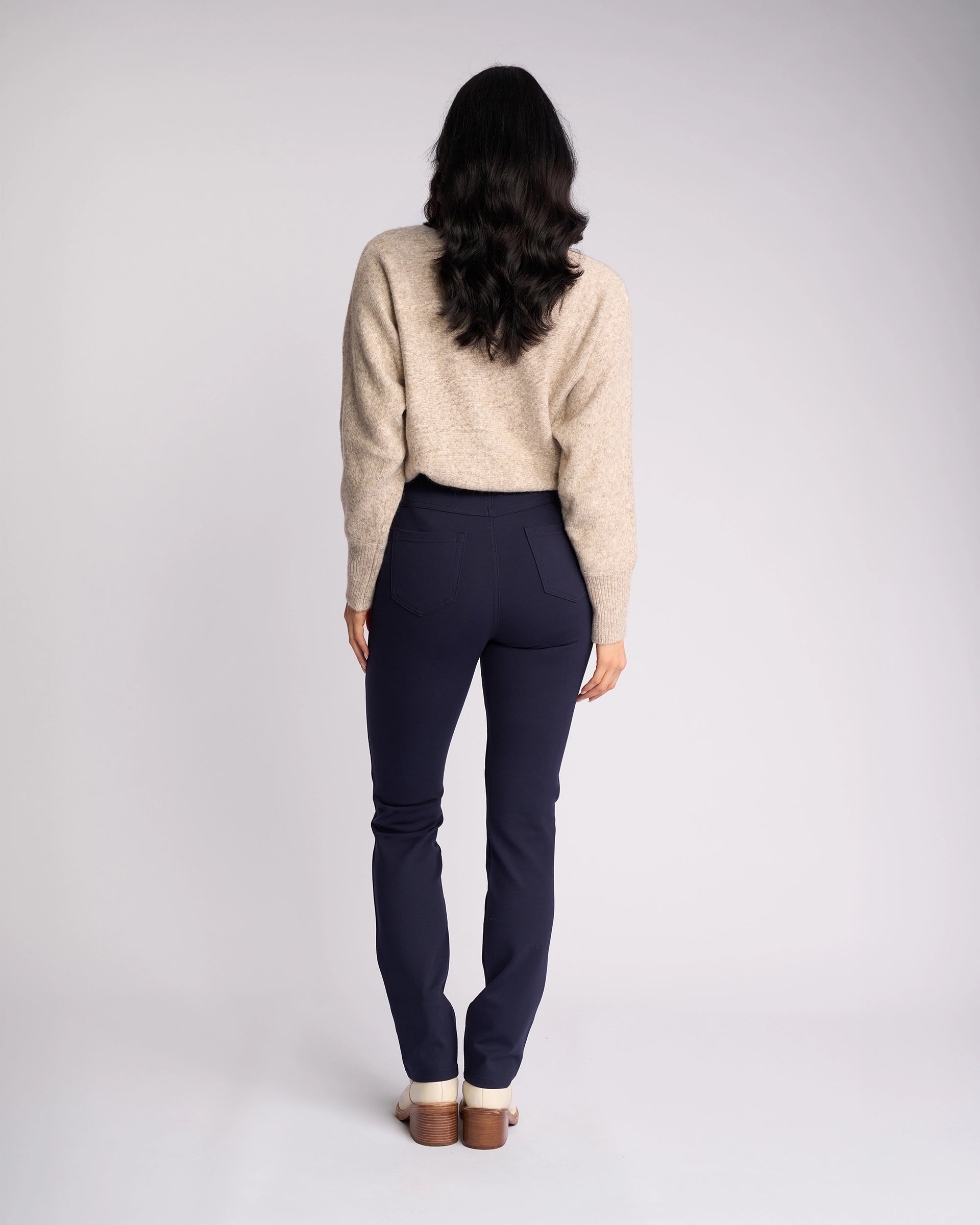 A person with long dark hair stands facing away, wearing a beige sweater, light shoes, and the Vassalli Bay Ponti Pull On pants in Navy with a slim-leg style, set against a plain light background.