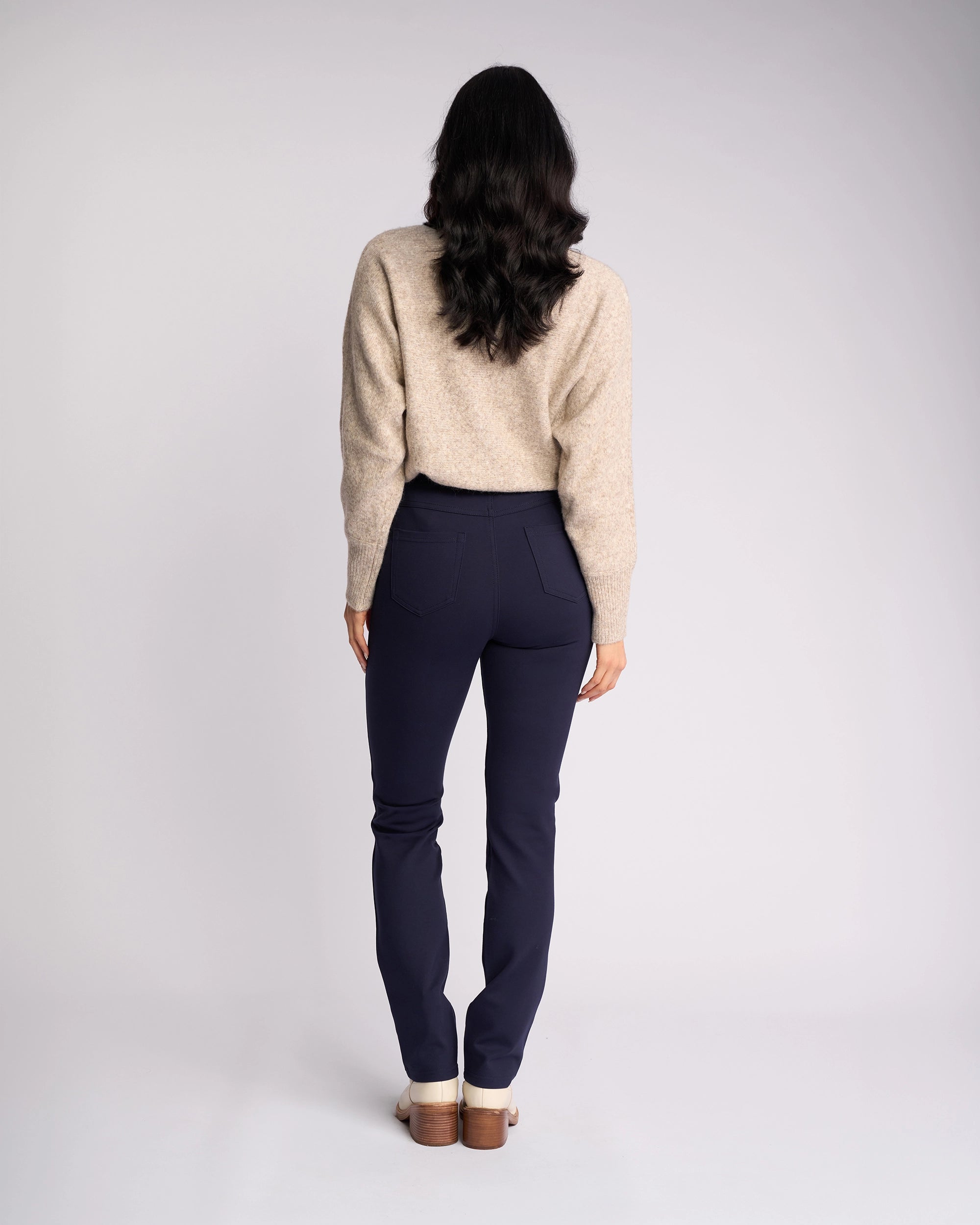 A person with long dark hair stands facing away, wearing a beige sweater, light shoes, and the Vassalli Bay Ponti Pull On pants in Navy with a slim-leg style, set against a plain light background.