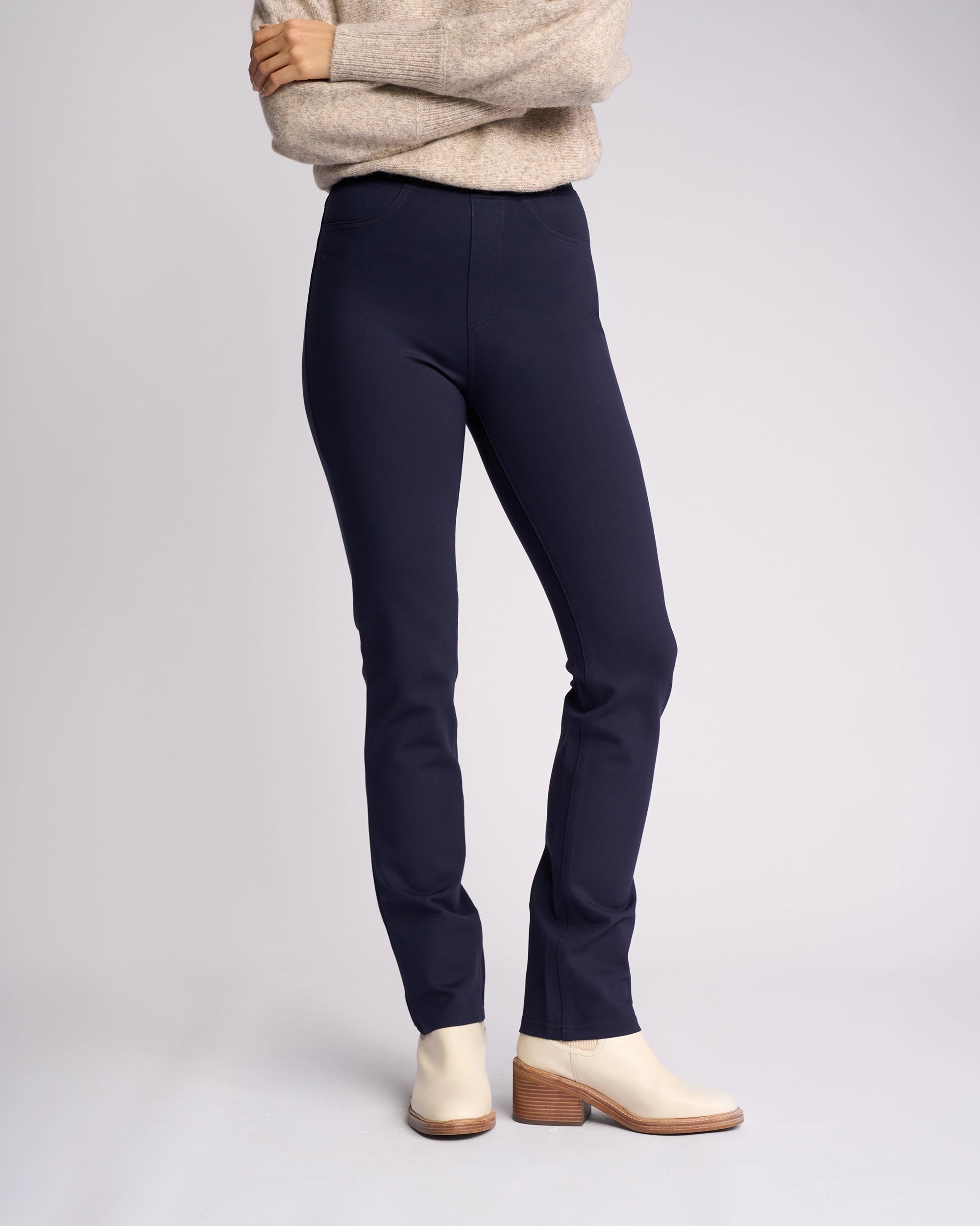 A person, arms folded and head not visible, stands against a plain light background wearing a beige sweater, cream ankle boots, and Vassalli's Bay Ponti Pull On - Navy pants for an effortless workwear style.