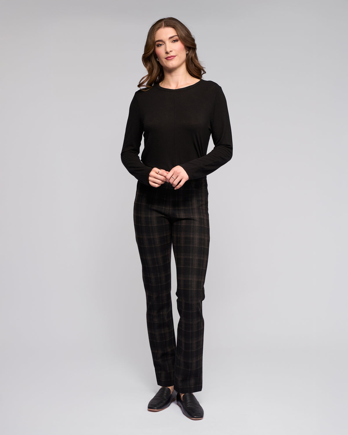 A person with long brown hair wears the Vassalli Bay Pull On - Checkmate slim pant and a black long-sleeve top, standing against a plain light gray background facing forward with hands loosely clasped.
