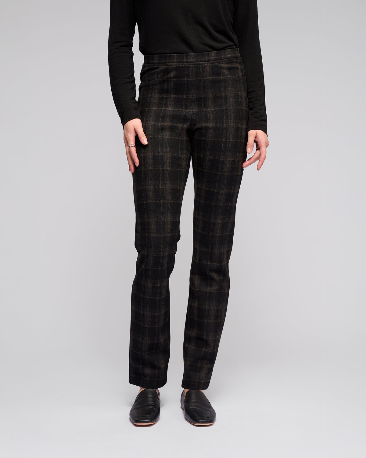 A person stands against a light gray background wearing Vassalli's Bay Pull On – Checkmate pants, a long-sleeve black top, and black slip-on shoes. The image is cropped from the shoulders down.