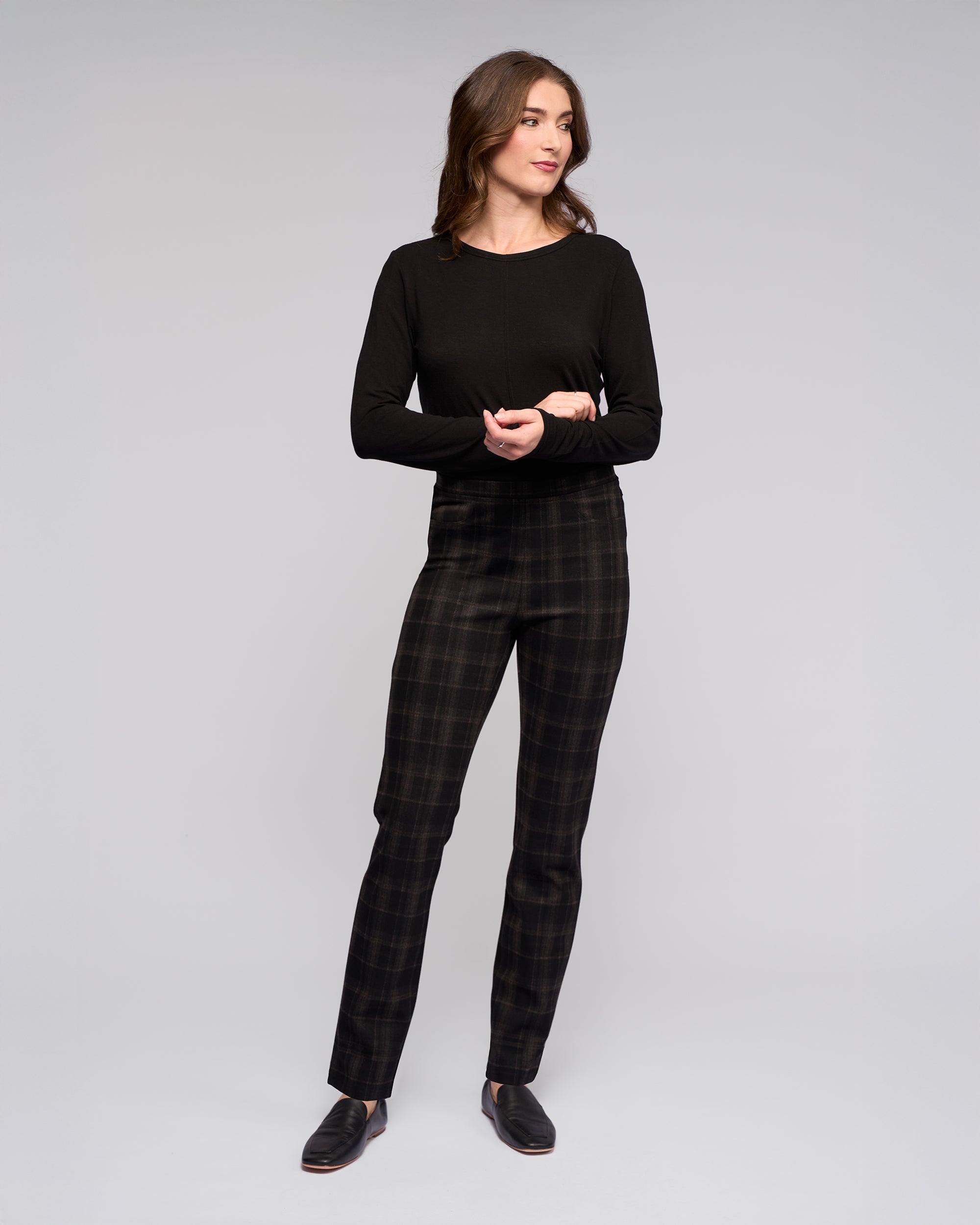 A woman stands against a plain light gray background, wearing Vassalli's Bay Pull On - Checkmate slim pants made from stretch ponti fabric, a black long-sleeve top, and black slip-on shoes. She looks to the side with a neutral expression, arms loosely crossed.