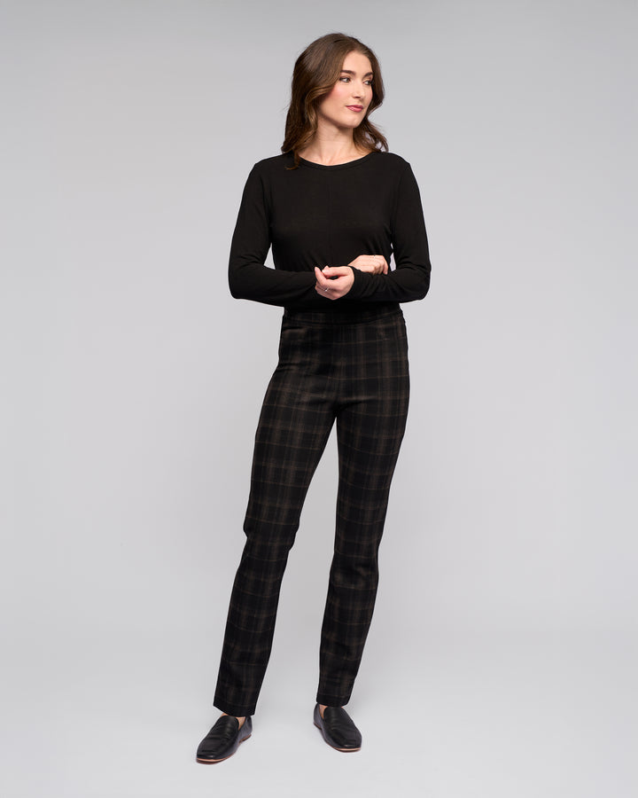A woman stands against a plain light gray background, wearing Vassalli's Bay Pull On - Checkmate slim pants made from stretch ponti fabric, a black long-sleeve top, and black slip-on shoes. She looks to the side with a neutral expression, arms loosely crossed.